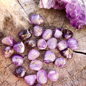 Puede incluir: Un conjunto de 20 piedras de runas de amatista con letras doradas sobre una superficie de madera. Las piedras están dispuestas en círculo con un gran cristal de amatista en el fondo.