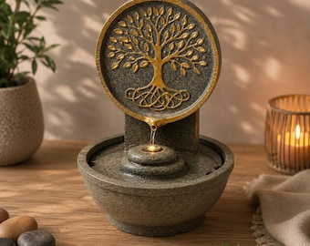 May include: A tabletop water fountain featuring a circular back panel with a gold-coloured tree of life relief. The fountain has a dark grey stone-like texture, a basin for water, and a small integrated light that illuminates the flowing water stream.
