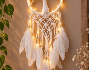 Könnte beinhalten: Ein weißer Makramee-Traumfänger mit einem zentralen Lebensbaum-Motiv aus Kordel und hellen Holzperlen. Der kreisförmige Rahmen ist mit warmen LED-Lichterketten beleuchtet, wobei weiße Federn an den unteren Strängen herabhängen.