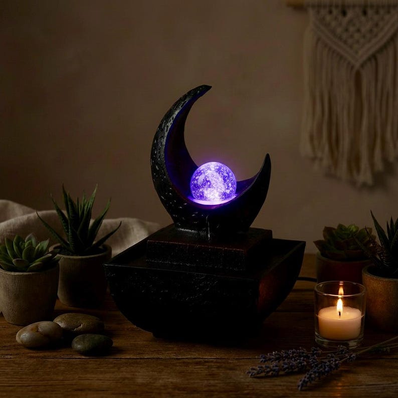 May include: A black water fountain with a crescent moon design, holding a glowing purple orb. The fountain sits on a wooden surface, surrounded by small potted plants, stones, and a lit candle. The overall theme is relaxation and home decor.