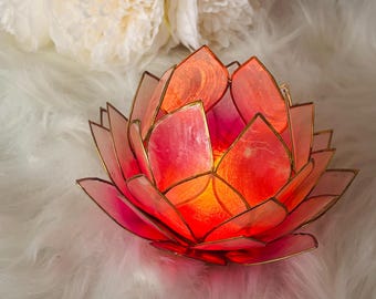 May include: A lotus flower candle holder crafted from translucent pink and orange capiz shell petals with gold-toned metal edging. The center glows with a warm light, highlighting the organic texture of the shell material in a layered design.