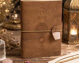 May include: A brown leather journal featuring embossed celestial designs: moons, sun, and geometric patterns. A rose quartz crystal is attached with a leather strap. The journal is closed with a string and rests on a wooden surface.