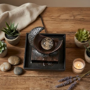 May include: A small, decorative water fountain with a crescent-shaped top and a glass orb. The fountain is black and bronze, with water gently flowing. Small potted succulents, stones, and a lit candle surround the fountain on a wooden surface.