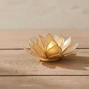 May include: A golden lotus flower-shaped candle holder. The petals are a light, translucent color, and the center is a solid gold. The candle holder sits on a round, gold base. The background is a light brown wooden surface.