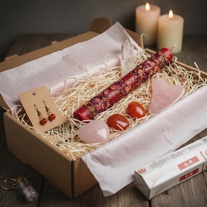 May include: A gift box filled with a red candle decorated with dried flowers, rose quartz stones, carnelian stones, earrings, and incense. The box is lined with paper and straw.