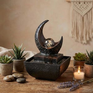 May include: A decorative indoor water fountain with a crescent moon design. The fountain features a textured, dark brown finish and a glass orb. The water flows from the orb into a square basin. Small potted succulents and a lit candle are nearby.