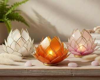 May include: Three lotus flower-shaped candle holders in white, orange, and pink. Each has a gold outline and a small candle inside, creating a warm glow. The candle holders are on a wooden surface.