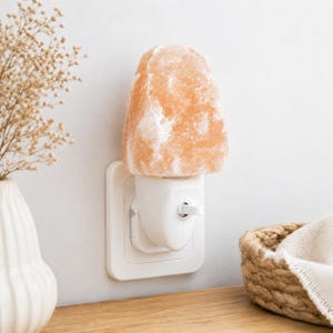 K&ouml;nnte beinhalten: Ein Himalaya-Salz-Nachtlicht, das in eine wei&szlig;e Steckdose gesteckt ist. Die Lampe hat einen rauen, orange-rosa Salzkristallschirm. Ein kleiner Korb und eine Vase mit getrockneten Blumen befinden sich im Hintergrund.