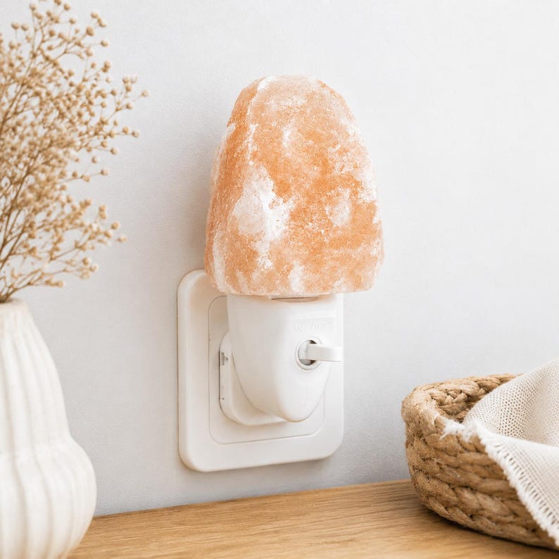 K&ouml;nnte beinhalten: Ein Himalaya-Salz-Nachtlicht, das in eine wei&szlig;e Steckdose gesteckt ist. Die Lampe hat einen rauen, orange-rosa Salzkristallschirm. Ein kleiner Korb und eine Vase mit getrockneten Blumen befinden sich im Hintergrund.