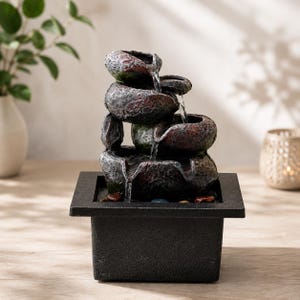 Puede incluir: Una fuente de agua de mesa de varios niveles con cuencos apilados de textura similar a la piedra en tonos carbón, óxido y verde musgo. El agua fluye desde el cuenco superior hacia abajo a través de los niveles hasta una base cuadrada negra mate.