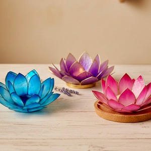 Può includere: Tre portacandele decorativi a forma di fiore di loto blu, viola e rosa. Il loto blu è a sinistra, il viola al centro e il rosa a destra. Ognuno ha un bordo dorato ed è realizzato in un materiale traslucido.
