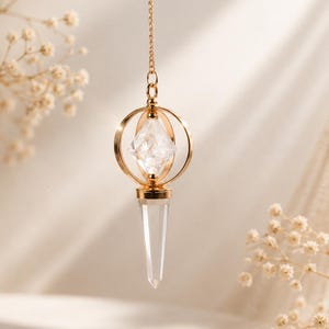 Könnte beinhalten: Ein goldfarbenes Metallpendel, das an einer feinen Kette hängt. Das Design besteht aus einem zentralen, klaren, facettierten Kristall in einem kreisförmigen Goldring, mit einer langen, spitzen, transparenten Kristallspitze an der Unterseite.