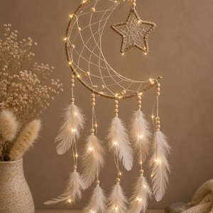 May include: A crescent moon and star shaped dreamcatcher wrapped in tan twine, featuring a woven web, light-colored wooden beads, and white feathers hanging from strings. The structure is illuminated by small, warm-toned LED fairy lights.