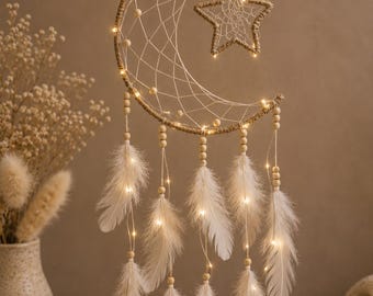 May include: A crescent moon and star shaped dreamcatcher wrapped in tan twine, featuring a woven web, light-coloured wooden beads, and white feathers hanging from strings. The structure is illuminated by small, warm-toned LED fairy lights.