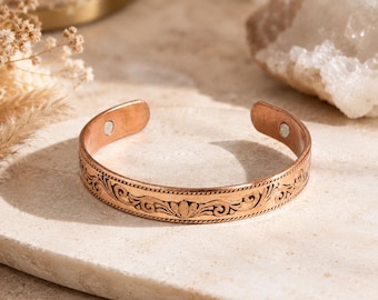 May include: A copper cuff bracelet featuring an intricate embossed floral scroll pattern along the outer band. The open-ended design includes two small circular magnets embedded on the interior ends of the metal band, providing a functional detail.