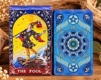 May include: A tarot deck box and a single card featuring a blue and yellow design. The box displays a figure walking on a cliff with a white dog under a yellow sun, labelled THE FOOL. The card back features a blue circular mandala pattern.