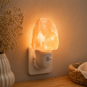 Puede incluir: Una luz de noche de sal del Himalaya enchufada a una toma de corriente. La lámpara tiene una pantalla de cristal de sal rugosa de color naranja-rosa que emite un brillo cálido. La toma de corriente y la base de la lámpara son blancas. El fondo es de color neutro.