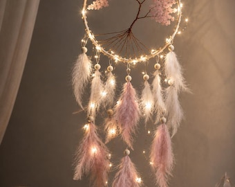 May include: A circular wall hanging featuring a wire tree sculpture with pink crystal leaves inside a hoop. The design includes white and light pink feathers suspended from the hoop, accented by small warm white LED fairy lights throughout.