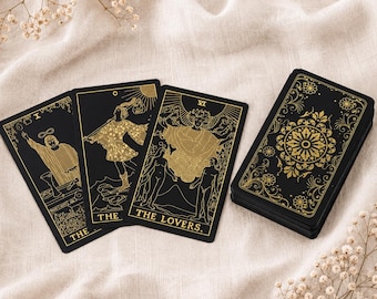 May include: A deck of tarot cards with black backgrounds and gold foil illustrations. Three cards are laid out face up, showing The Magician, The Fool, and The Lovers, while a stack of cards sits to the right featuring an ornate gold floral pattern.