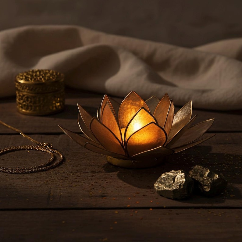 May include: A golden lotus-shaped candle holder with a lit candle inside. A small, ornate golden box and a delicate gold necklace are also visible. Two pieces of pyrite are on the wooden surface.