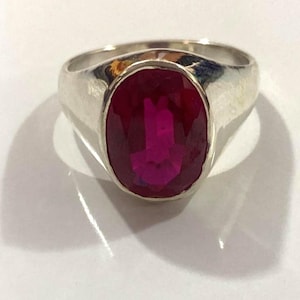 Peut inclure: Une bague en argent avec une grosse pierre précieuse rouge foncé de forme ovale. La pierre précieuse est l'élément central, sertie dans un anneau en argent poli simple. La bague est présentée sur une surface blanche, avec une ombre sur le côté.