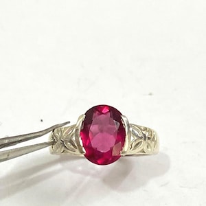 May include: A silver ring featuring an oval, deep red gemstone. The ring's band has a floral design on the sides. The gemstone is held in place by four prongs. The ring is set against a white background.