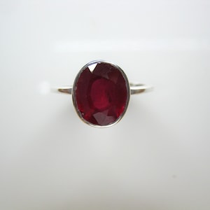May include: A silver ring featuring a large, oval-cut, deep red gemstone. The gemstone is the focal point, set in a simple, elegant band. The ring is displayed against a white background.