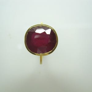 May include: A gold ring featuring a large, faceted, oval-shaped ruby gemstone. The ruby is a deep, rich red color, set in a gold bezel. The ring's band is a simple, thin gold design.