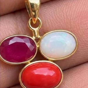 May include: A gold pendant featuring three oval-shaped stones arranged in a cluster. The pendant includes one faceted deep red ruby, one smooth white opal with iridescent flashes, and one smooth bright red coral stone, all set in polished gold.