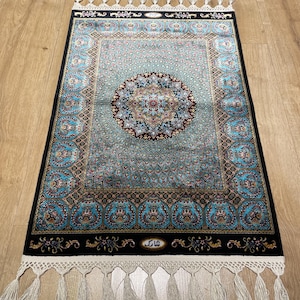 Può includere: Un tappeto rettangolare con un design a medaglione centrale nei toni del turchese, blu e rosso. Ha un bordo nero con motivi floreali e nappe bianche. Il tappeto è posato su un pavimento in legno. Dimensioni: circa 100 cm x 60 cm.