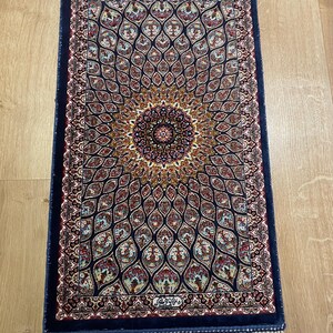 Op de afbeelding: Een rechthoekig, sierlijk tapijt met een marineblauwe rand en een centraal medaillonontwerp. Het tapijt heeft een gedetailleerd patroon in tinten rood, goud en blauw, met een crèmekleurige franje aan de korte uiteinden. Het tapijt is waarschijnlijk gemaakt van geweven materiaal.