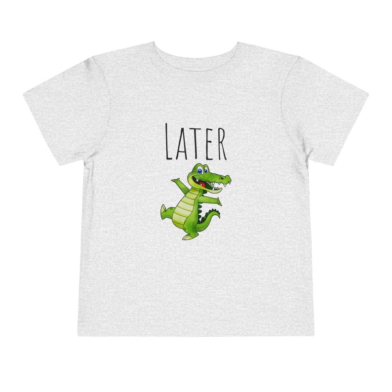 K&ouml;nnte beinhalten: Ein heidefarbenes T-Shirt mit einem Comic-Krokodil und dem Wort "LATER" auf der Vorderseite.