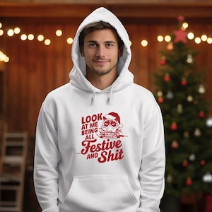 Puede incluir: Sudadera con capucha blanca con un gráfico rojo de un personaje parecido a Papá Noel y el texto "Look at me being all Festive and Shit". La sudadera tiene un bolsillo delantero y una capucha con cordón.