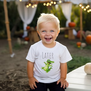 K&ouml;nnte beinhalten: Ein kleiner Junge tr&auml;gt ein wei&szlig;es T-Shirt mit einem gr&uuml;nen Alligator-Motiv und dem Wort "LATER" darauf. Er l&auml;chelt und schaut in die Kamera.