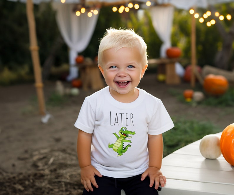 K&ouml;nnte beinhalten: Ein kleiner Junge tr&auml;gt ein wei&szlig;es T-Shirt mit einem gr&uuml;nen Alligator-Motiv und dem Wort "LATER" darauf. Er l&auml;chelt und schaut in die Kamera.