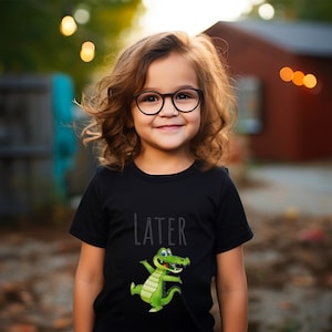 K&ouml;nnte beinhalten: Ein junges Kind tr&auml;gt ein schwarzes T-Shirt mit einem gr&uuml;nen Alligator-Motiv und dem Wort "LATER" darauf. Das Kind l&auml;chelt und tr&auml;gt eine Brille.