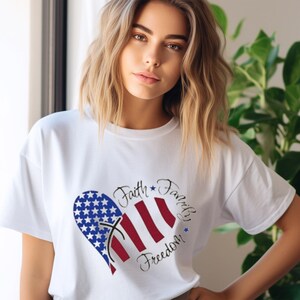 Puede incluir: Camiseta blanca con un dise&ntilde;o de coraz&oacute;n rojo, blanco y azul. El coraz&oacute;n est&aacute; formado por una bandera estadounidense y tiene una cruz en el centro. El texto "Faith Family Freedom" est&aacute; escrito alrededor del coraz&oacute;n.