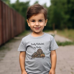 May include: A young child wearing a gray t-shirt with the text "Easily Distracted by Rocks." The shirt features a graphic of rocks and foliage. The child is smiling, with a blurred green background.