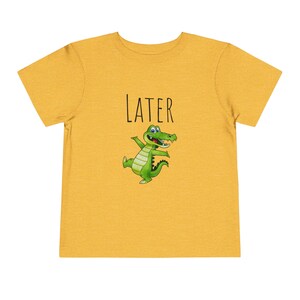 K&ouml;nnte beinhalten: Ein gelbes T-Shirt mit einem gr&uuml;nen Alligator-Cartoon und dem Wort "LATER" in schwarzem Text.