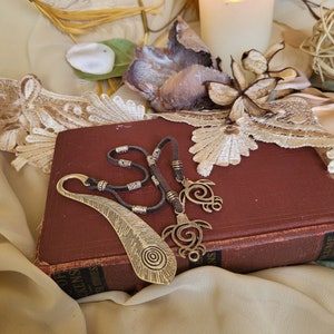 May include: A still-life image features a dark red book with a gold feather-shaped bookmark and a necklace with turtle pendants. A lit white candle and dried floral elements are in the background. The necklace is made of dark gray cord.