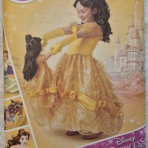 Puede incluir: Un sobre de patrón de costura Simplicity 8407 para un disfraz de Bella en tallas 3-8. La portada muestra un vestido amarillo con falda de capas, guantes a juego, una versión para muñeca del vestido y la marca Disney Princess.