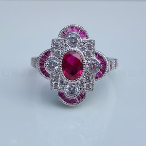 May include: A silver ring with a central oval ruby surrounded by white and pink gemstones. The ring has an Art Deco design with a symmetrical pattern of stones and a detailed band. The gemstones are set in a silver metal.