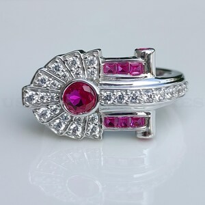 May include: A silver ring with a large pink ruby stone surrounded by smaller white stones. The ring has a geometric design with three rows of smaller pink stones.
