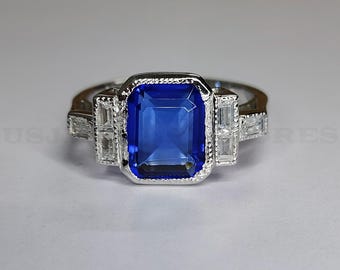 May include: A silver ring featuring a large, rectangular, deep blue gemstone as the centerpiece. The ring is accented with smaller, rectangular, clear gemstones on either side of the main stone. The band has a detailed design.