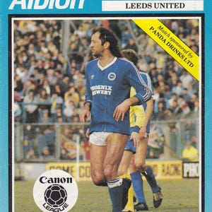 May include: A vintage soccer program cover featuring the Brighton & Hove Albion team. The cover includes the text "Brighton & Hove Albion" in white letters on a blue background. The image shows a soccer player in a blue jersey with white shorts. The program is from the 1984-85 season.