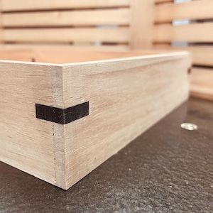 May include: Close-up of a light-colored wooden box with a black corner joint. The box is rectangular and appears to be handmade. The wood grain is visible. The box is likely for storage or display.