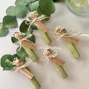 May include: Five small glass vials filled with green powder, each tied with twine and a small bouquet of white flowers. A brown paper tag is attached to each vial with the text "Eda & Samed" and the date "01.12.2023". The text "Minima Atölye" is written on the table.