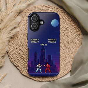 May include: A phone case featuring a pixelated video game scene. The design shows two characters in a fighting stance against a cityscape backdrop under a starry night sky. The screen displays scores and a timer.