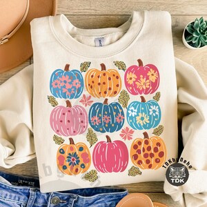 May include: Cream-colored sweatshirt featuring a grid of nine colorful pumpkins. Each pumpkin is a different color, including blue, orange, pink, and yellow, and decorated with floral patterns. The design is perfect for fall.