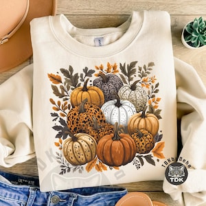May include: Cream-colored sweatshirt featuring a colorful pumpkin design. The graphic includes various pumpkins in shades of orange, white, and brown, surrounded by fall foliage. A perfect autumn-themed apparel item.